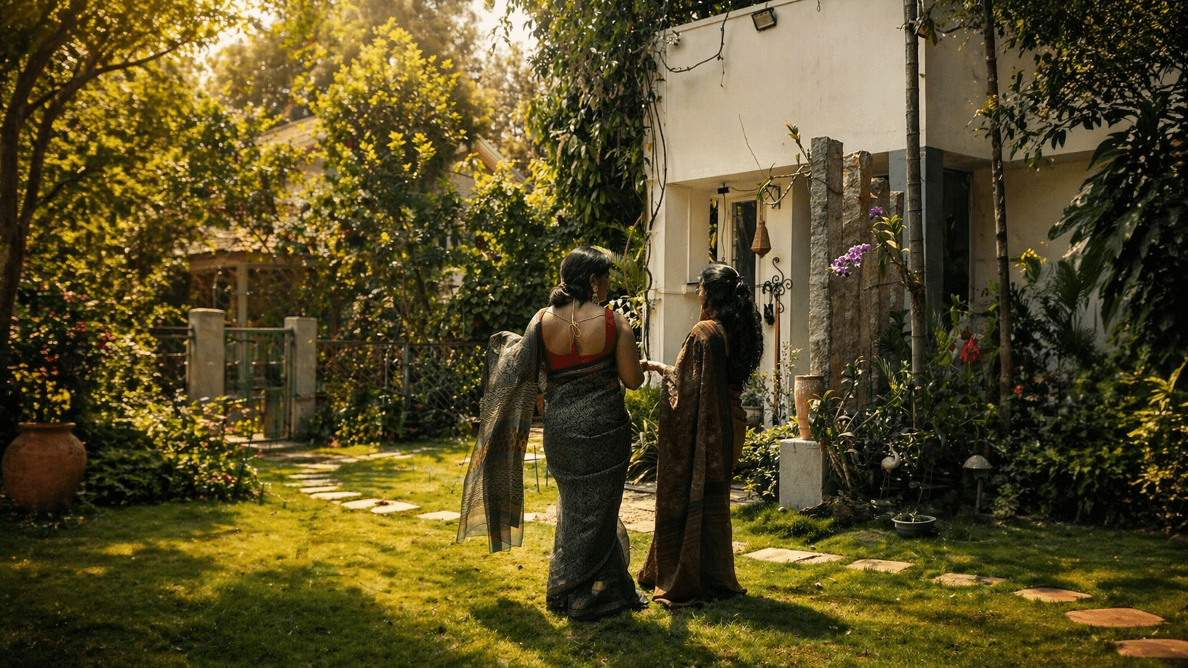 Two women in sarees walking through a sunlit garden